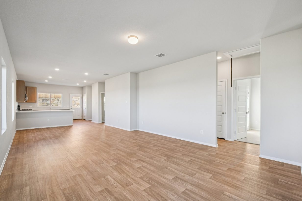 Image of a living room with light grey walls, vinyl flooring, and a kitchen in the distance