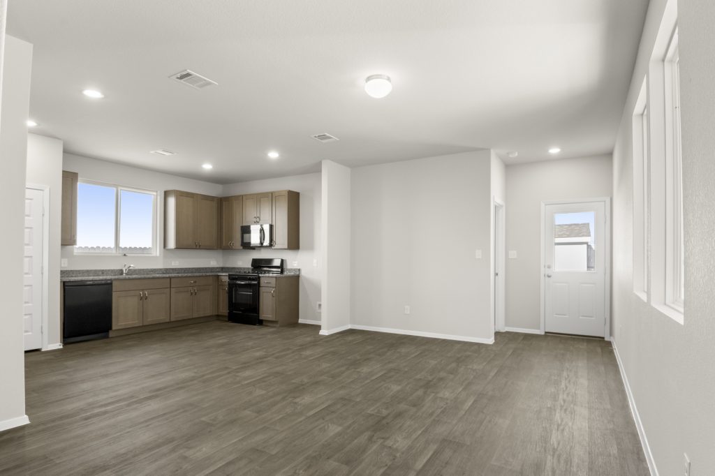 Image of a living room area with brown vinyl flooring and cream walls with a L-shaped kitchen in the corner