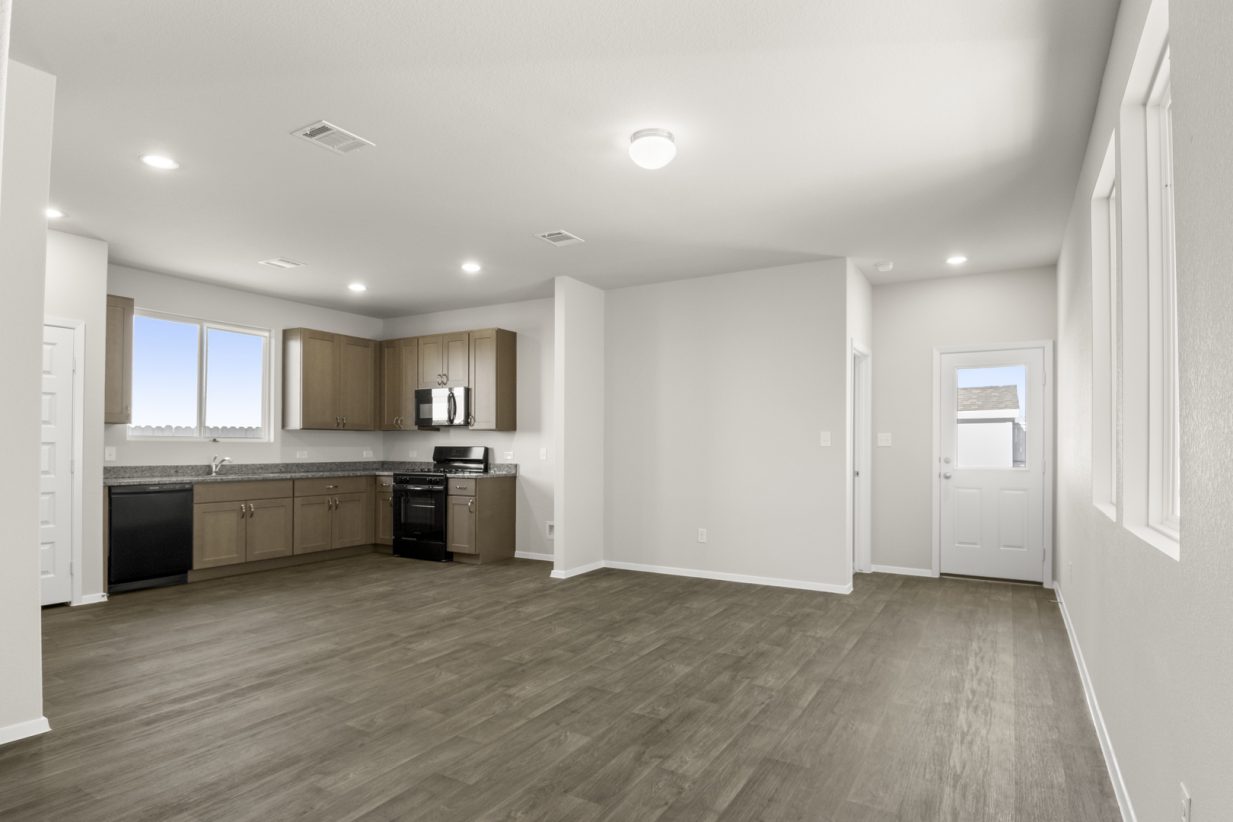 Image of a living room area with brown vinyl flooring and cream walls with a L-shaped kitchen in the corner