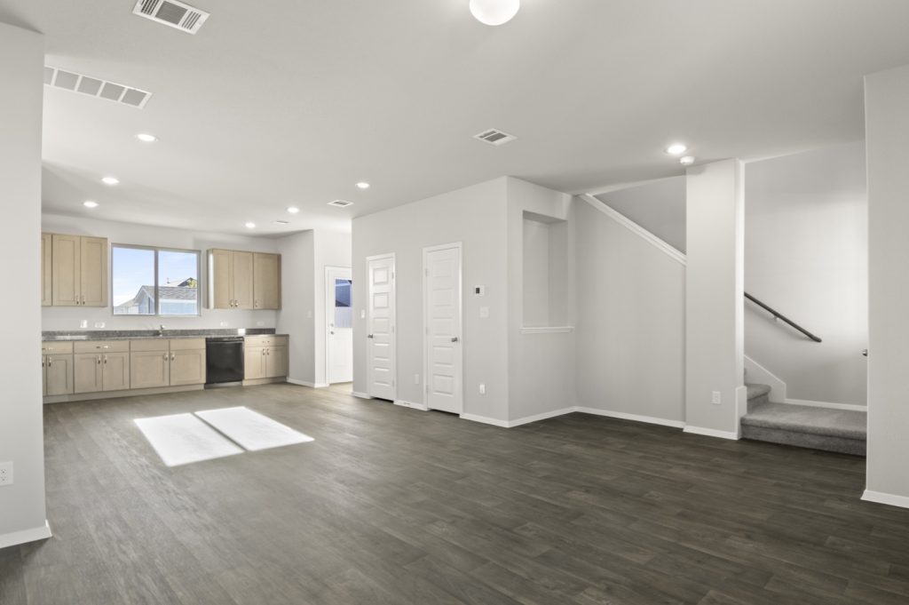 Image of a living room with dark vinyl flooring, light grey walls, a staircase that leads upstairs and a connecting open kitchen