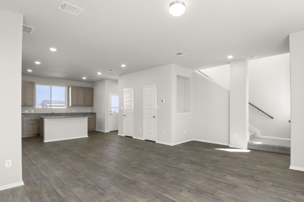 Image of a living room with light grey walls, a staircase, dark brown vinyl flooring, and a kitchen in the distance