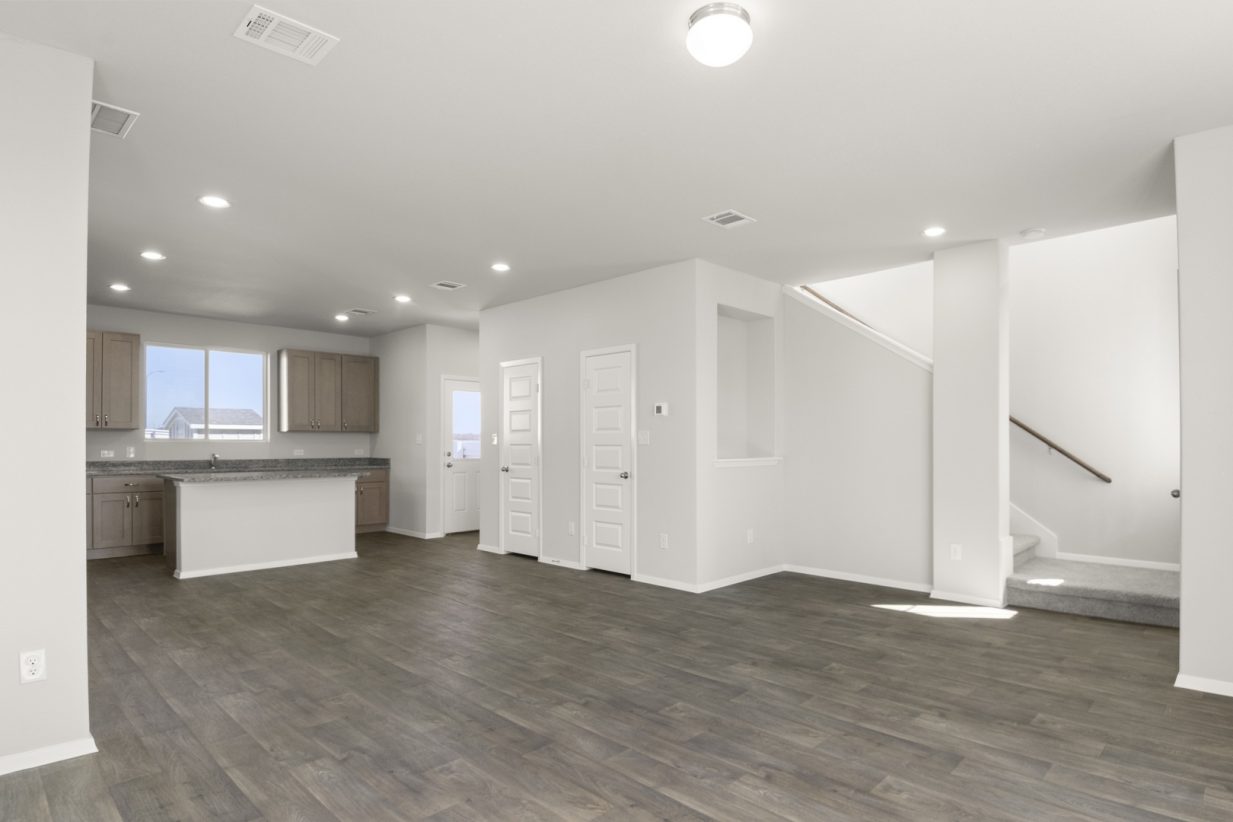 Image of a living room with light grey walls, a staircase, dark brown vinyl flooring, and a kitchen in the distance