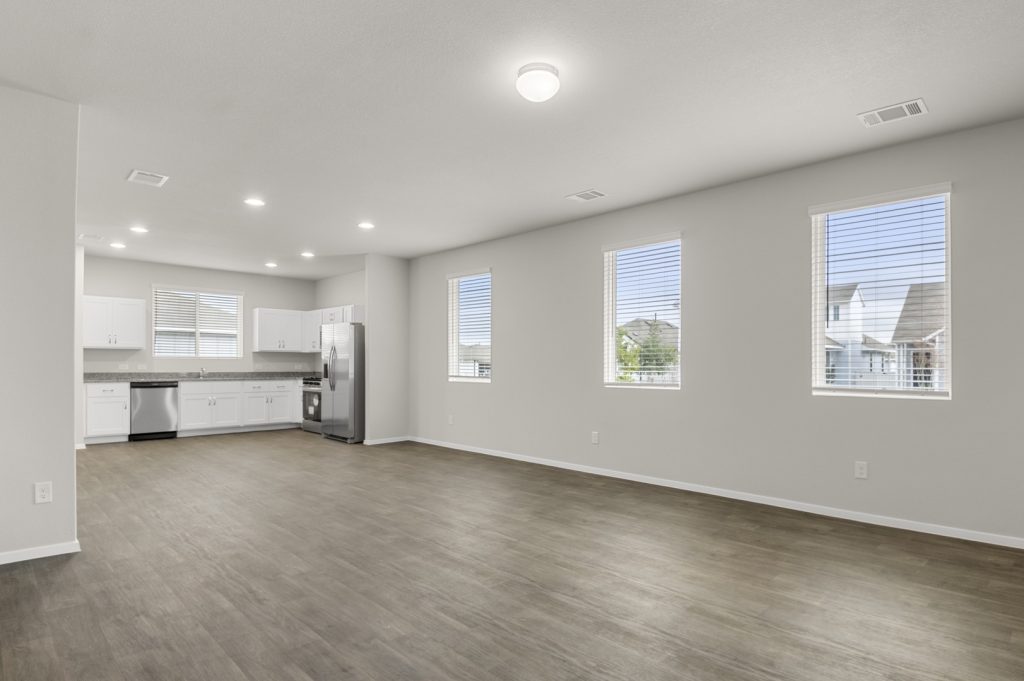 Image of a one-story home living room with brown vinyl flooring, light grey walls, three windows, and a kitchen in the distance