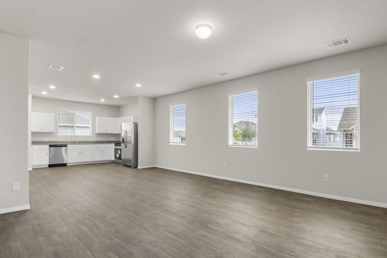 Image of a one-story home living room with brown vinyl flooring, light grey walls, three windows, and a kitchen in the distance