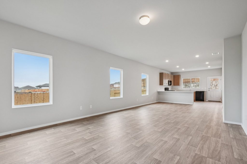 Image of a living room with light grey walls, vinyl flooring, windows, and a kitchen in the distance