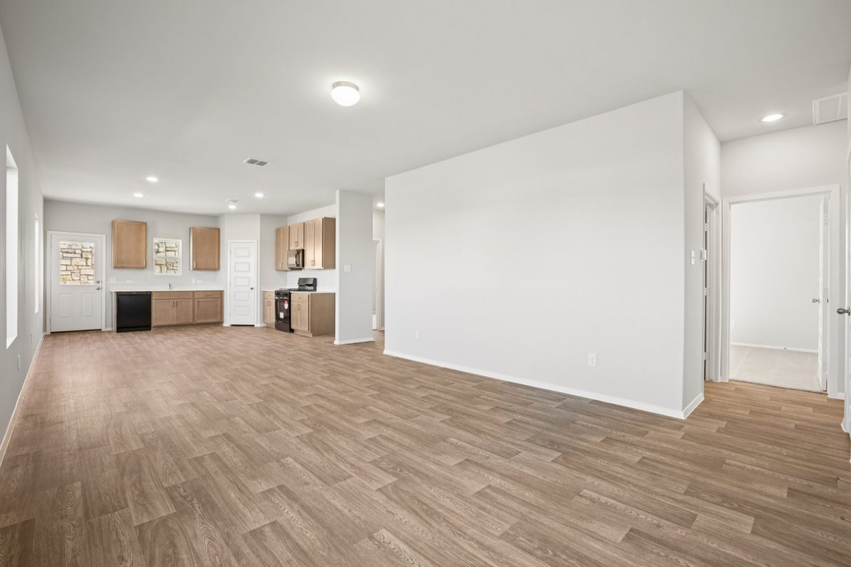 Image of a living room area with light grey walls, vinyl flooring, a kitchen in the distance and white trim