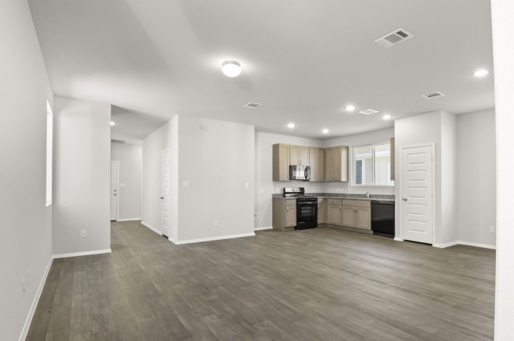 Image of a living room with dark vinyl flooring, light grey walls, a left front door pathway and a connecting L-shaped kitchen