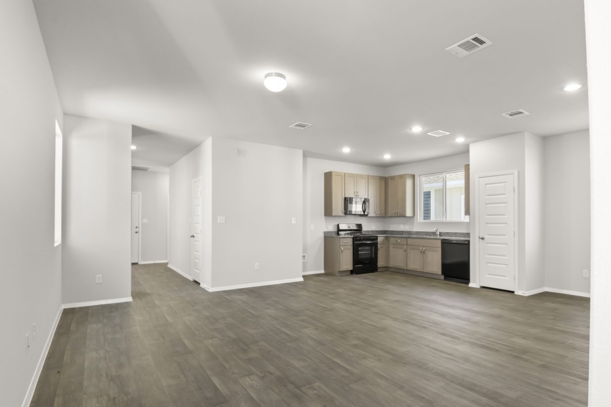 Image of a living room with dark vinyl flooring, light grey walls, a left front door pathway and a connecting L-shaped kitchen