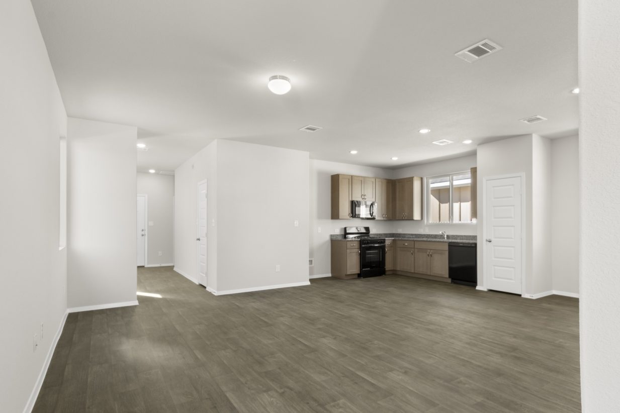Image of a living room with light grey walls, dark brown vinyl flooring, an L-shaped kitchen in the distance and white trim