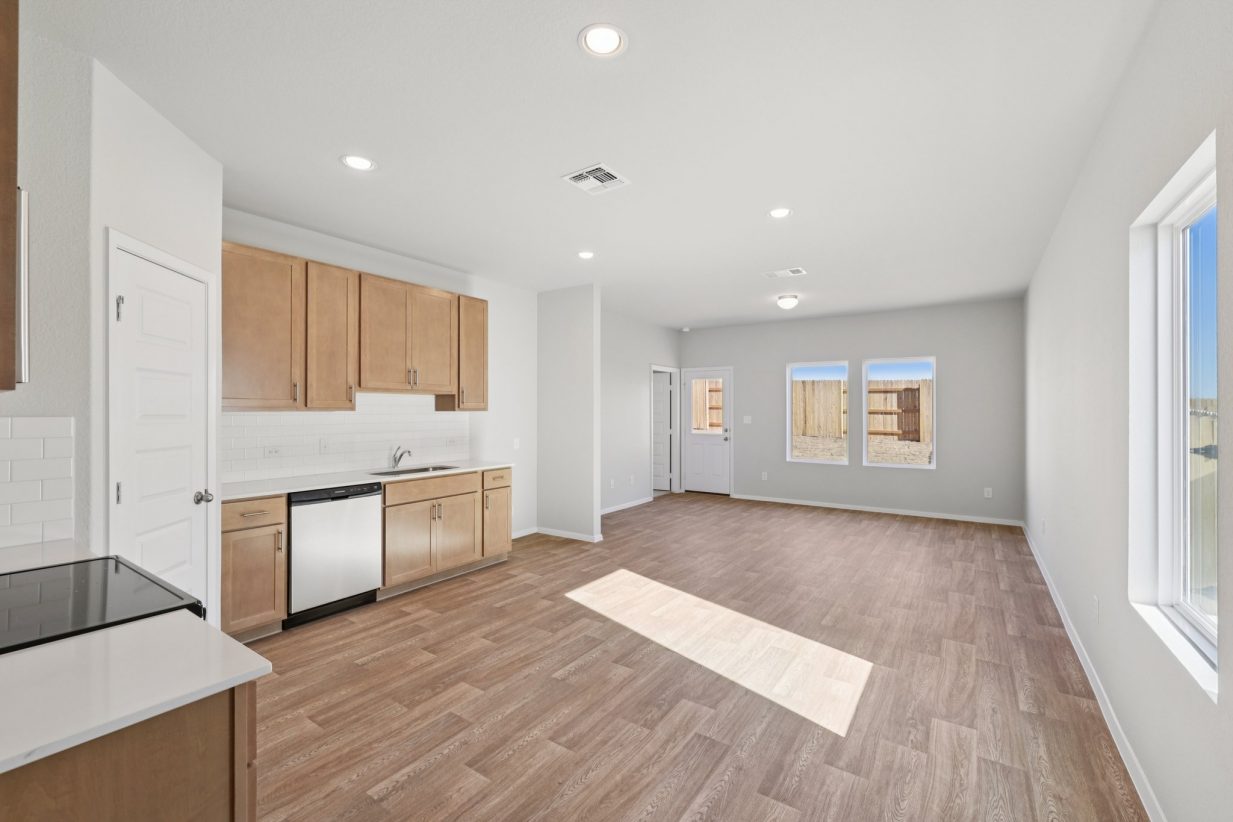 Image of a living room with light grey walls, brown wood-look vinyl flooring, windows and a kitchen with light brown cabinets