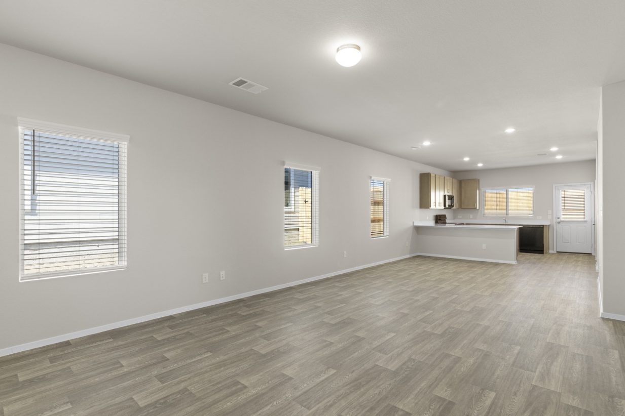 Image of a living room with beige walls, light brown wood-look flooring, windows with blinds, and an open kitchen in the distance