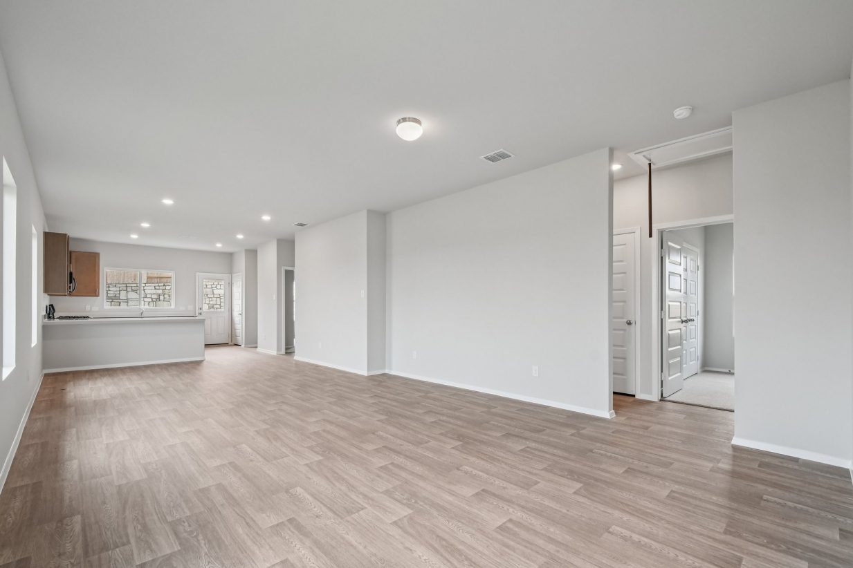 Image of a living room with light grey walls, vinyl flooring and a kitchen in the distance