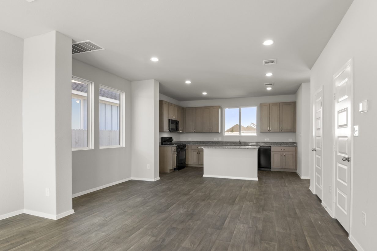 Image of a dining room with light grey walls, dark brown vinyl flooring, a dining room area, and a kitchen in the distance