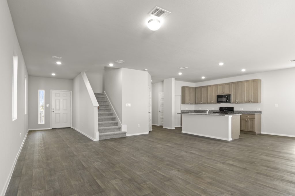 Image of a living room with a grey carpeted staircase leading upstairs and a kitchen with a center island in the corner