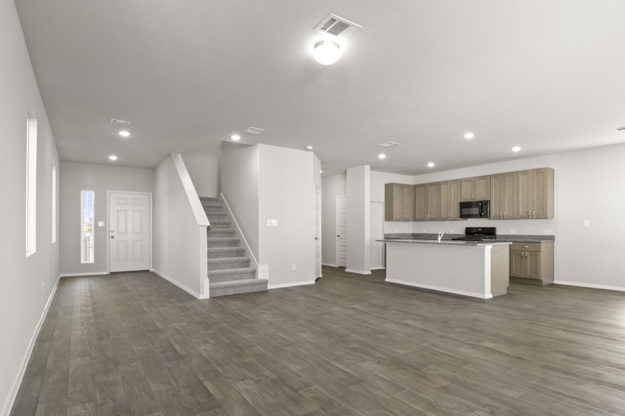 Image of a living room with a grey carpeted staircase leading upstairs and a kitchen with a center island in the corner