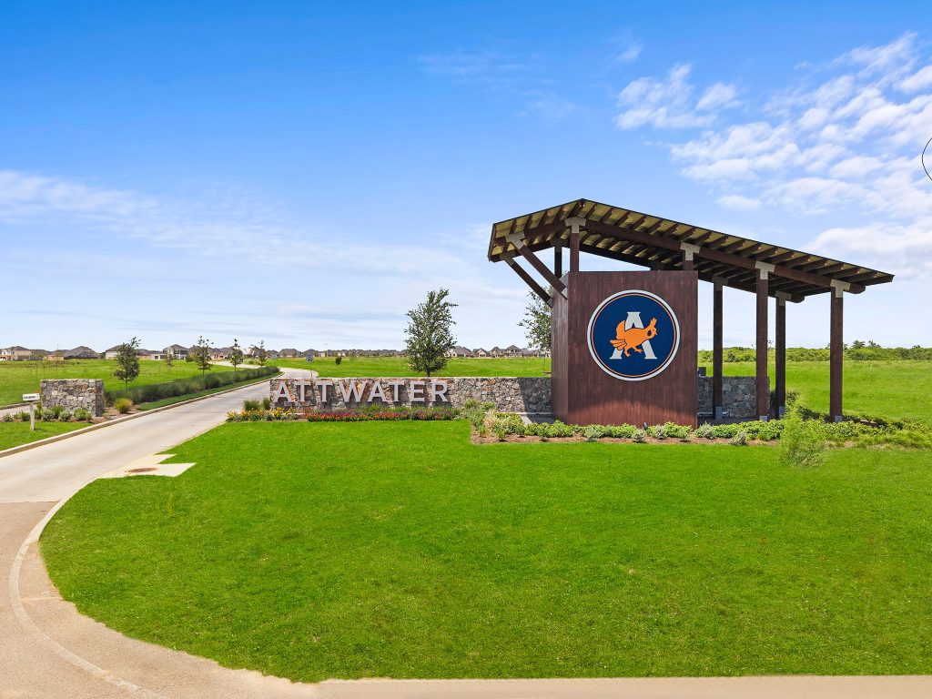 Entrance monument for the Attwater community featuring a large wooden and stone sign with the Attwater name and logo, surrounded by manicured landscaping, open green space, and a roadway leading into the neighborhood under a bright blue sky.