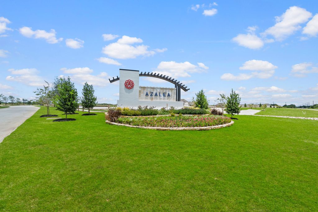 Entrance monument for the Azalea community featuring a stone sign with the Azalea name and orange logo, surrounded by manicured landscaping, open green space, and a roadway leading into the neighborhood under a bright blue sky with white clouds.