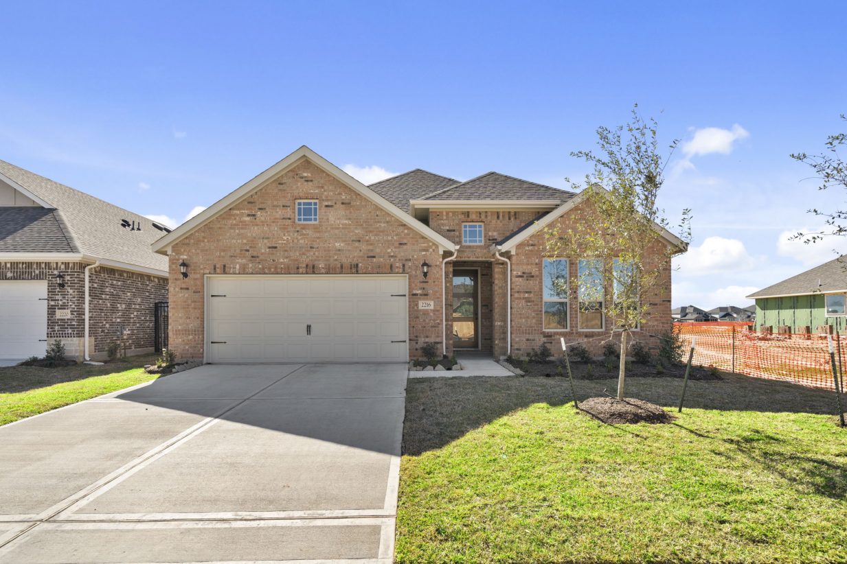 Image of a one story brick out with a white garage door, a cement driveway and green grass with a blue sky in the background