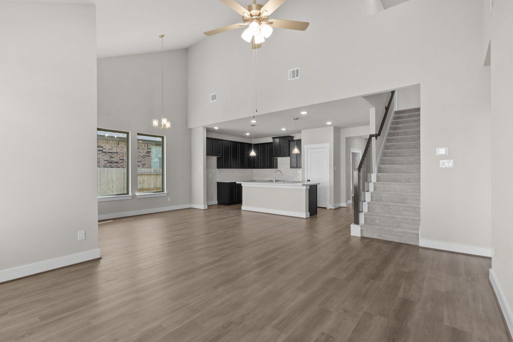 Image of a living room with beige walls, light brown wood-look flooring, carpeted stairs, and an open kitchen with a center island