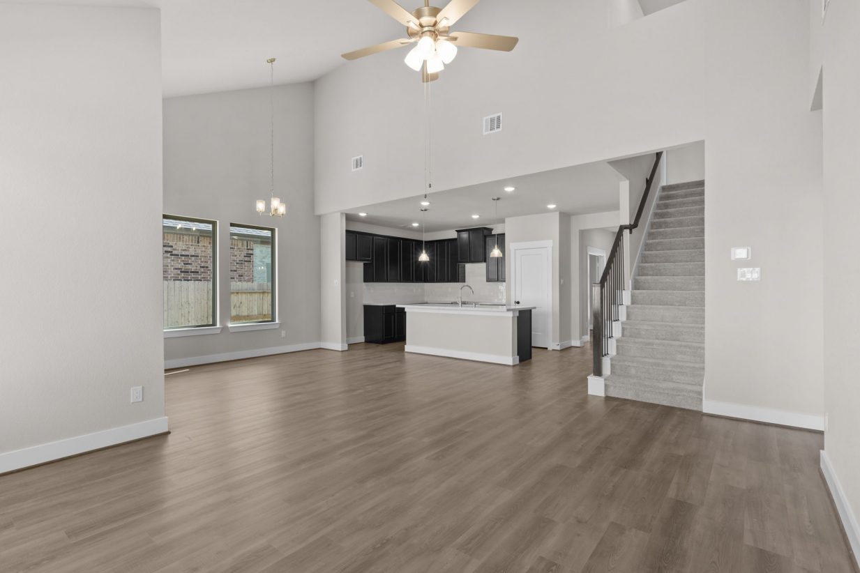 Image of a living room with beige walls, light brown wood-look flooring, carpeted stairs, and an open kitchen with a center island