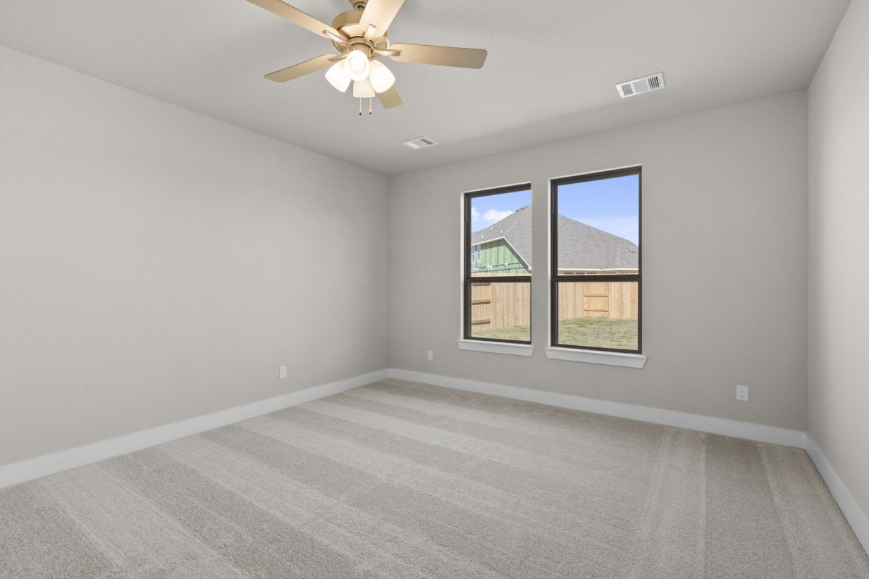 Image of a bedroom with beige walls, tan carpeting, two windows with black trim and a ceiling fan