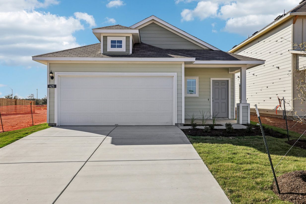Image of a green one story house with a grey front door, cement driveway, a white garage and a blue sky in the background