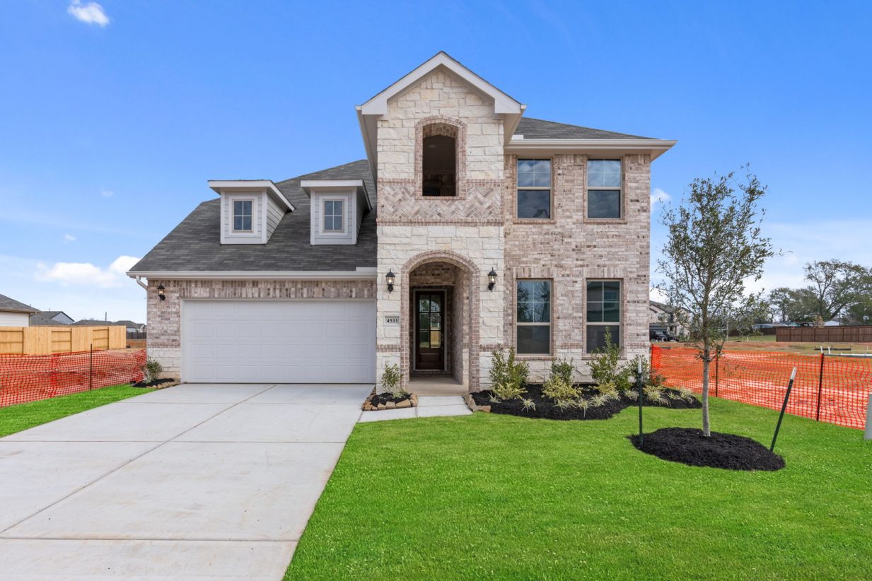 Image of a brick two story home with a white garage, cement driveway, green grass and a blue sky in the background