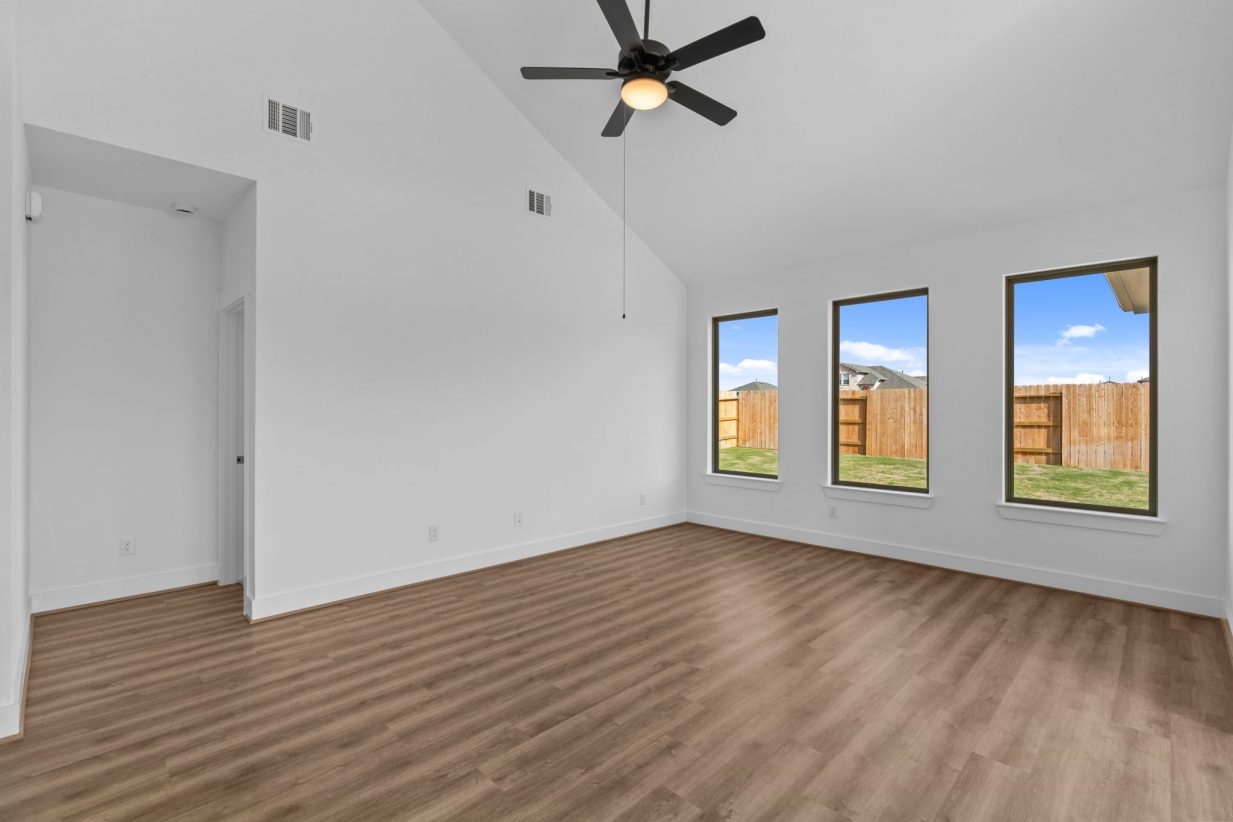Image of a living room with white walls, brown wood-look flooring, windows with brown trim, and a ceiling fan