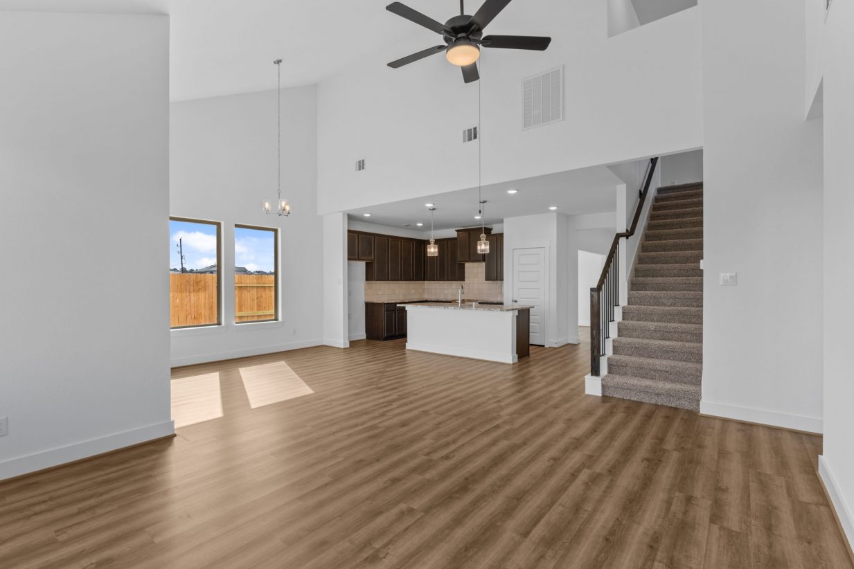 Image of a the interior of a two story house with white walls, brown wood-look flooring, with a kitchen and dining room open to living area, and a brown carpeted staircase