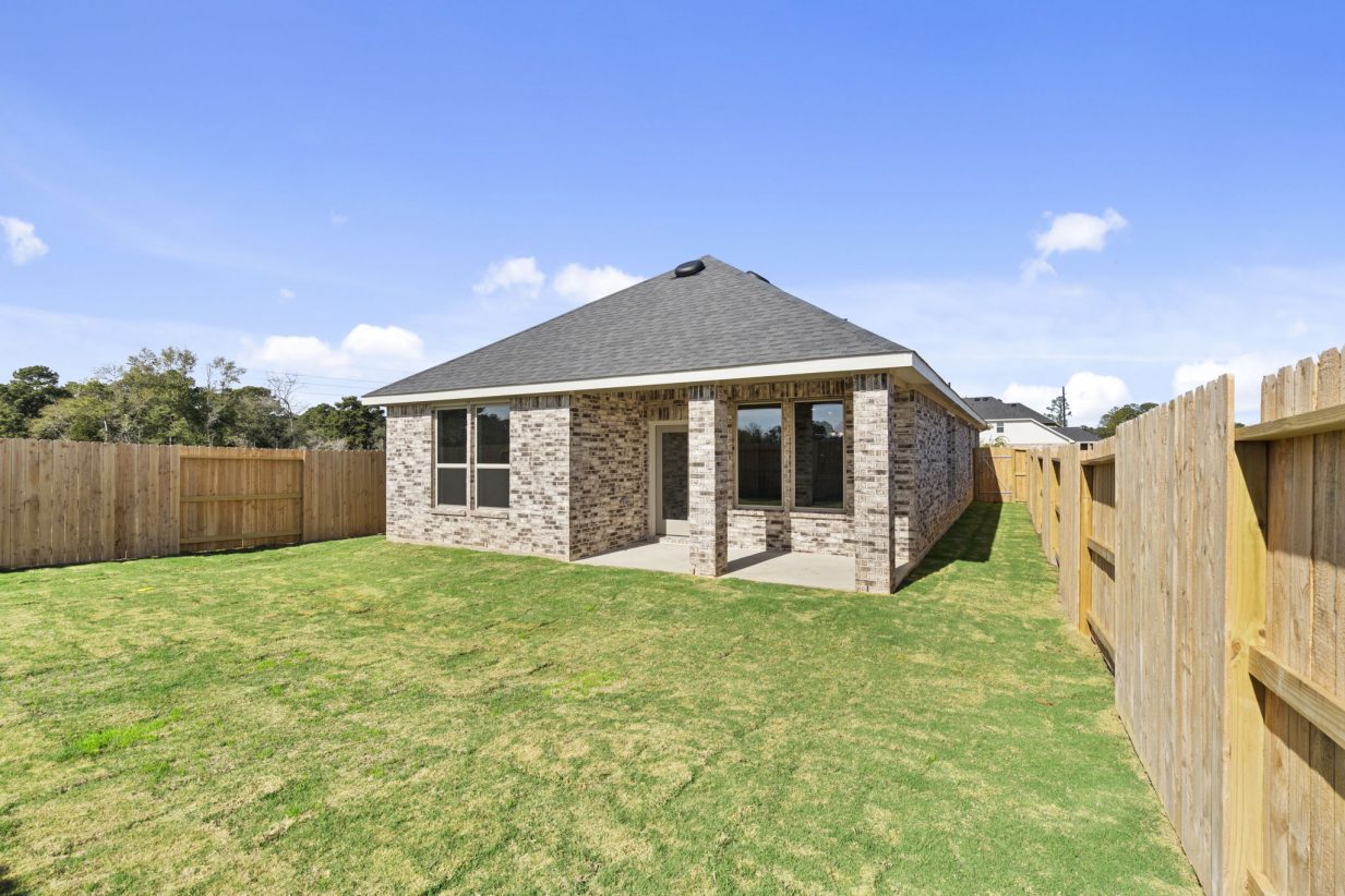 Image of the back exterior of a brick one story house with a covered back patio, green grass backyard, a wooden fence and a blue sky in the background
