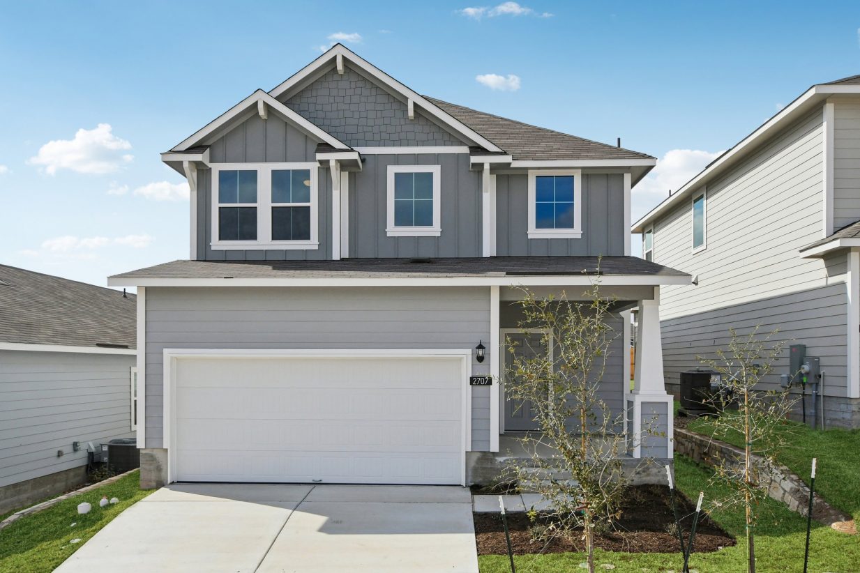 Image of a grey two story house front exterior with a white garage, a green grass front yard, and a blue sky in the background