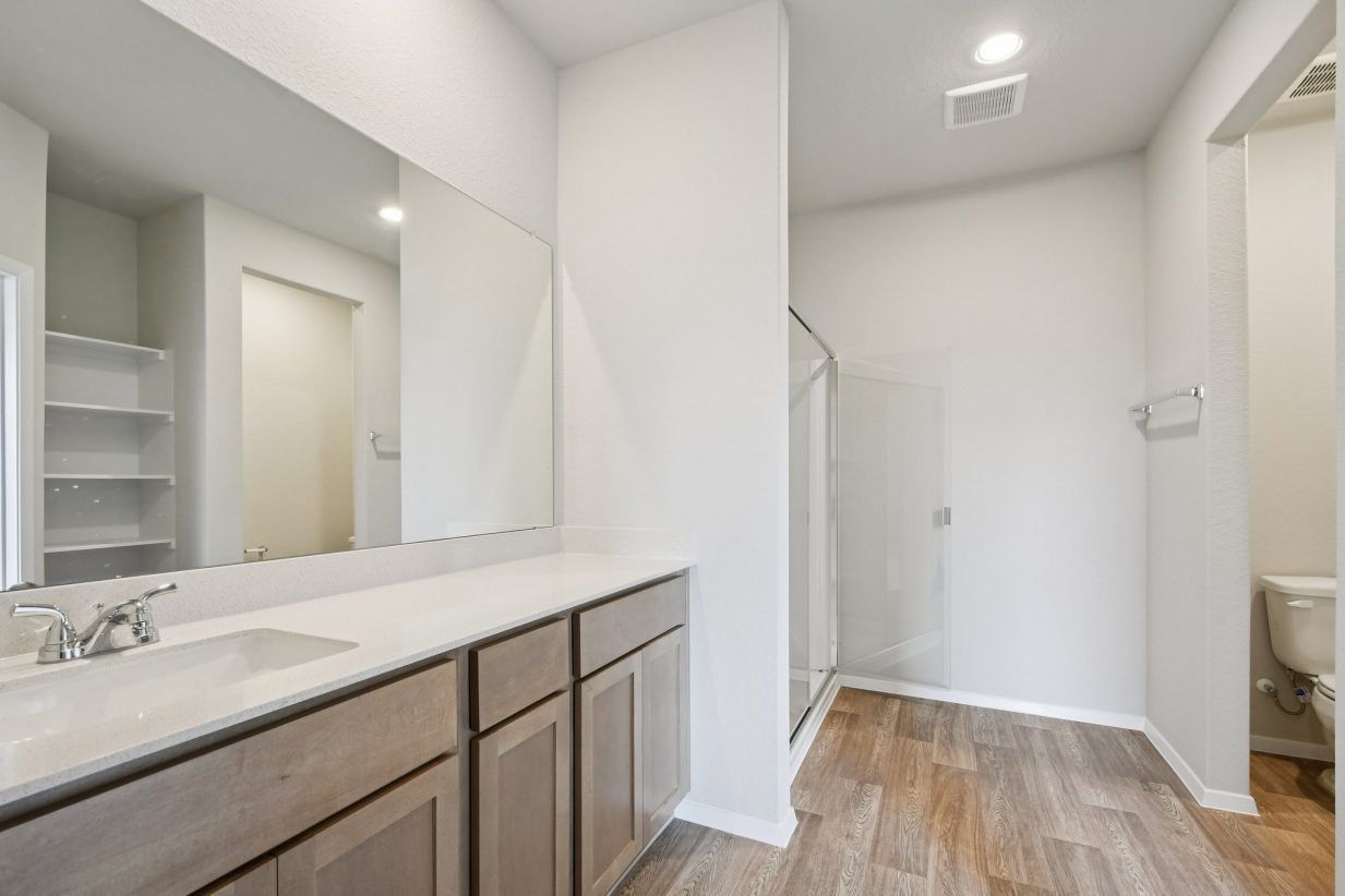 Image of a bathroom with light grey walls, brown cabinets, a white dual vanity, and a large mirror