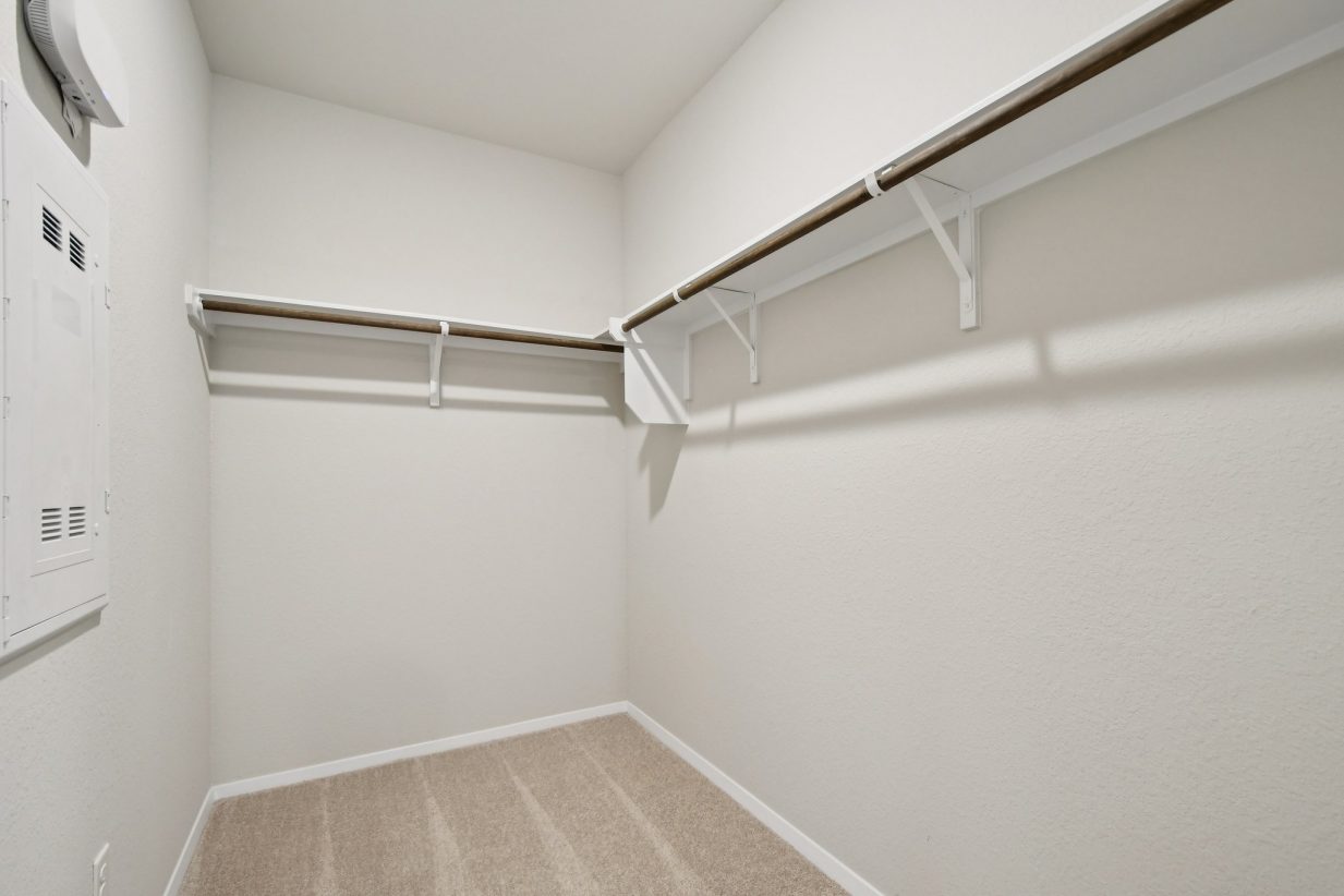 Image of a primary closet with light grey walls, tan carpeting, and wooden rods