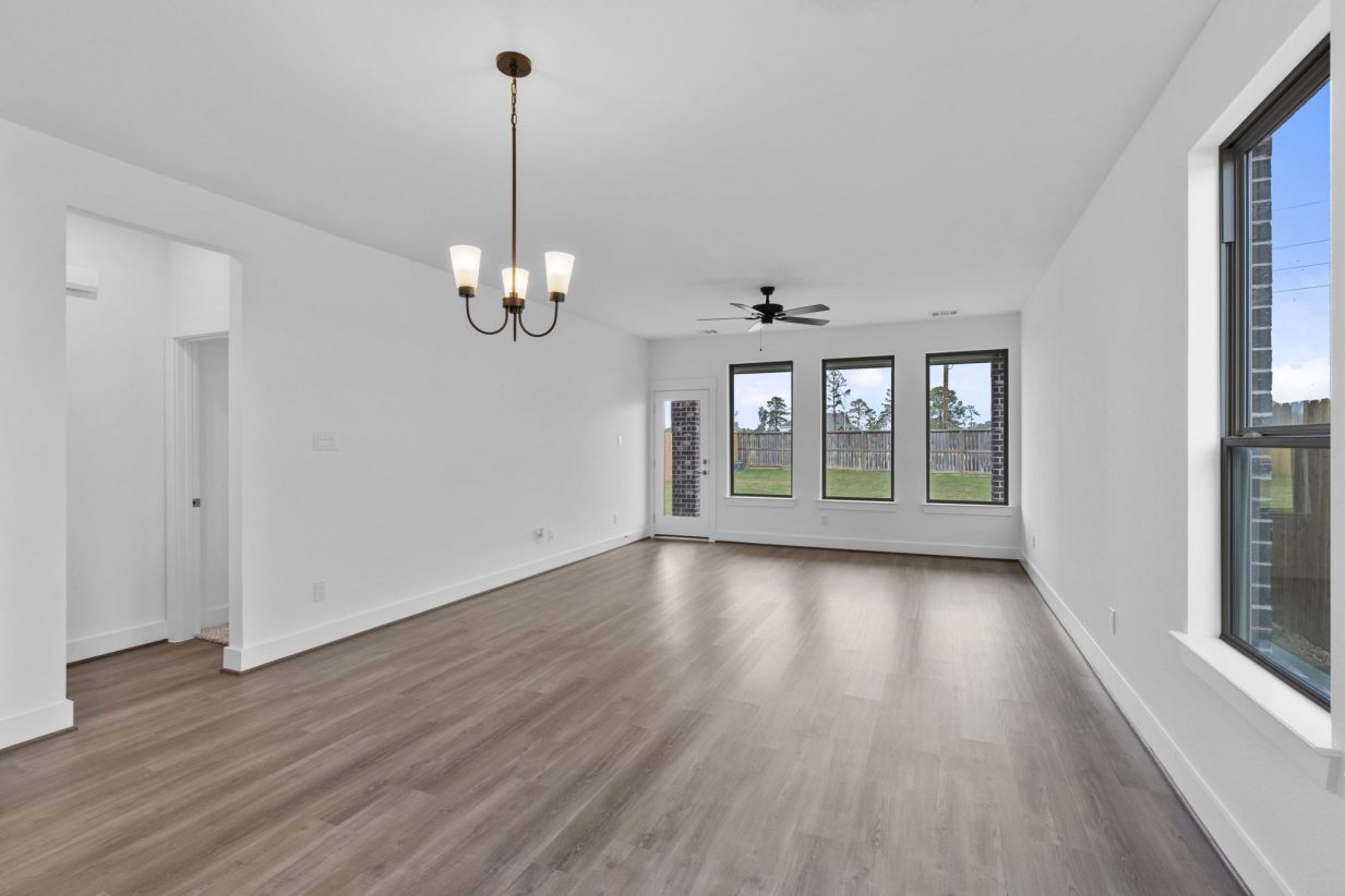 Image of a long living room with white walls, three windows with black trim, light brown wood-look flooring and a ceiling fan