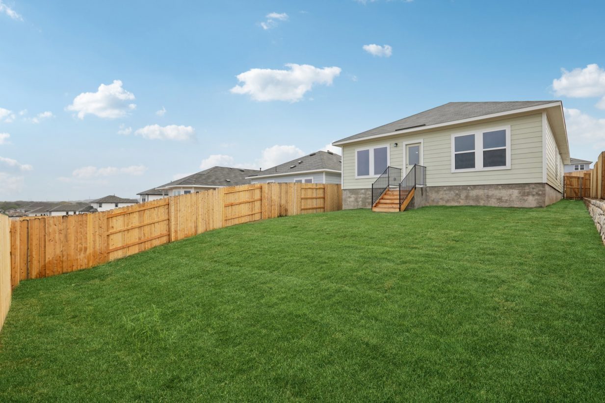 Image of a green one story house with a green grass backyard, a wooden fence and a blue sky in the background