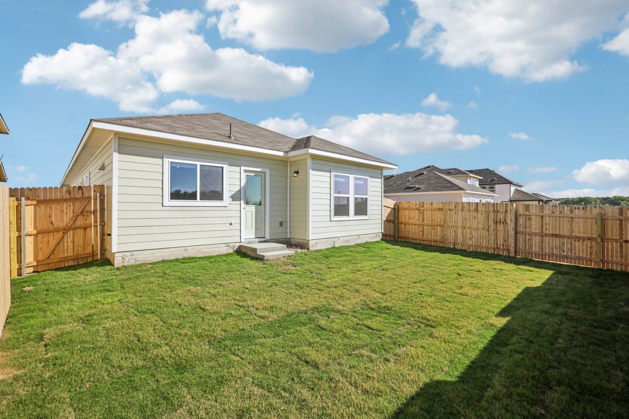 Image of a one story green house back exterior with green grass, a wooden fence and a blue sky in the background