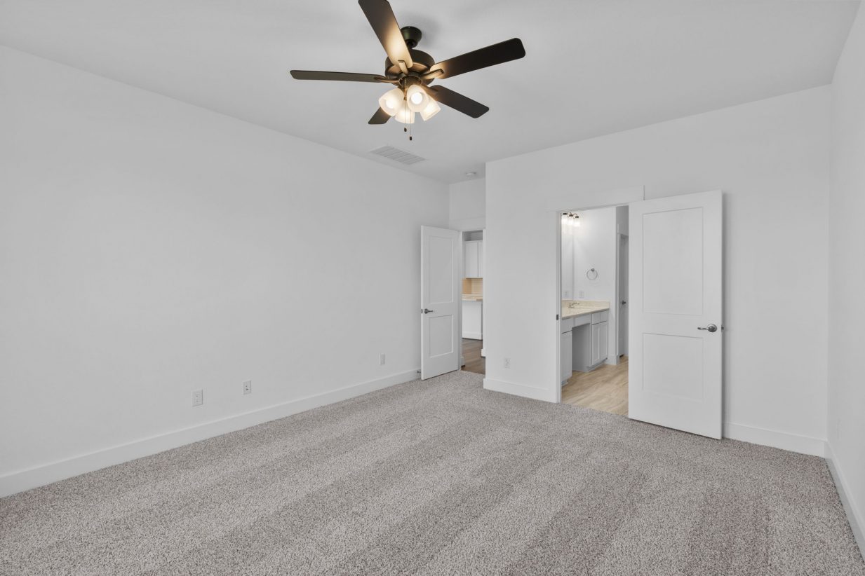Image of a primary bedroom with tan carpeting, white walls, and a ceiling fan
