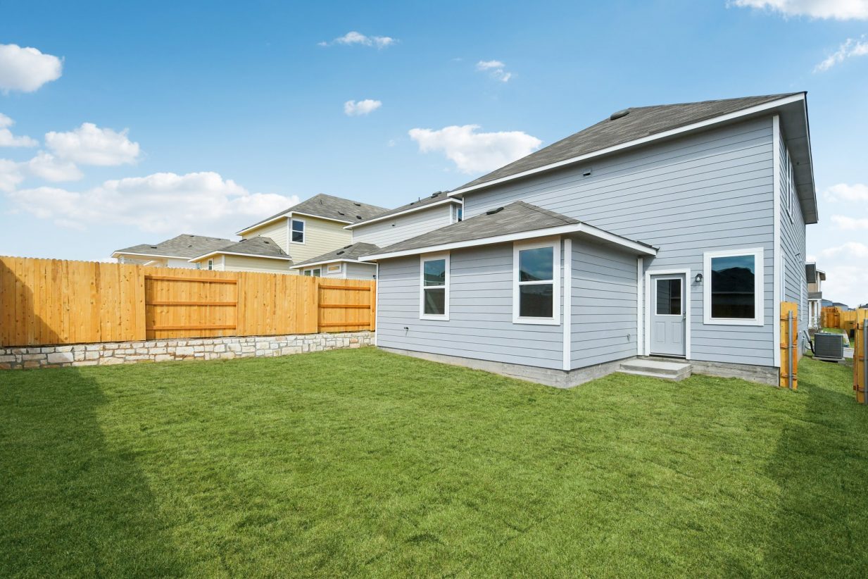 Image of the back exterior of a grey two story house with a green grass backyard, a wooden fence and a blue sky in the background