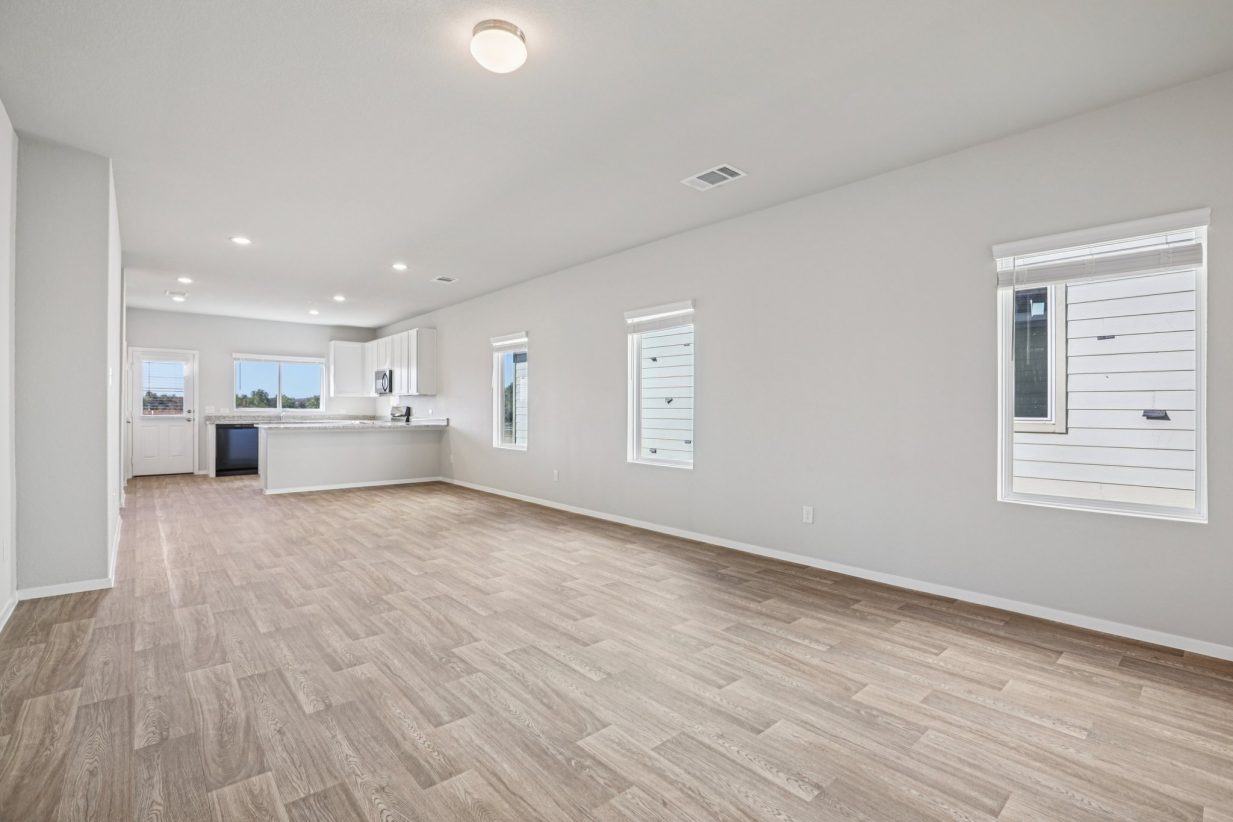 Image of a living room area with grey walls, vinyl flooring, windows and a kitchen in the distance
