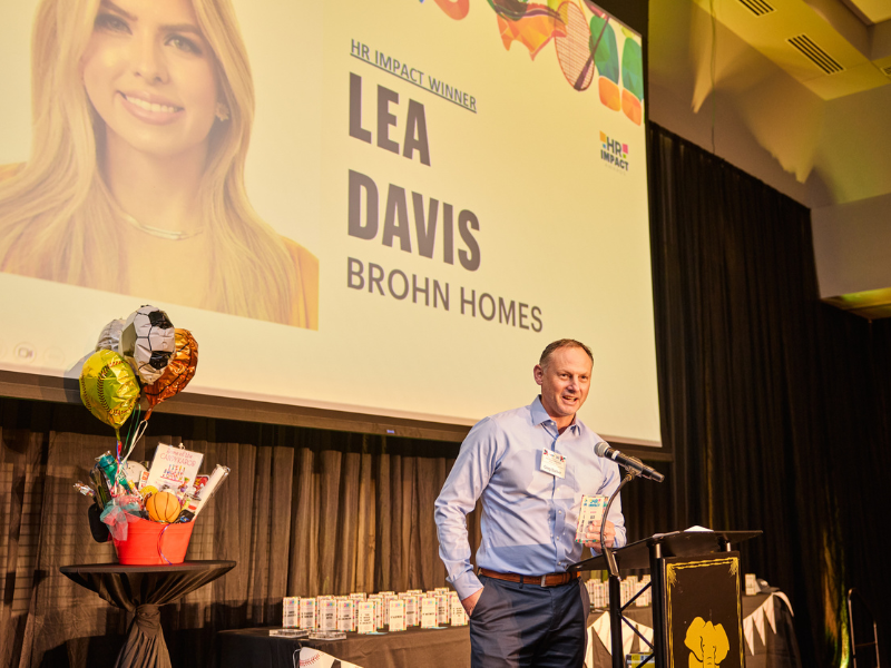 An image of a man in a blue long sleeve shirt in front of podium on a stage with a projection screen with the words "Lea Davis" on it.