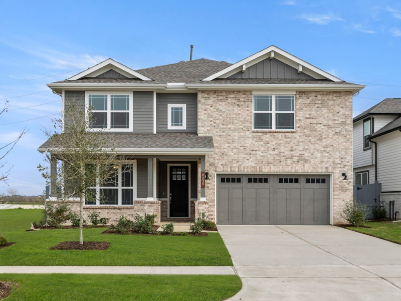 An image of two-story home with gray garage, white framed windows, green grass lawn with a tree, a concrete driveway, and blue sky background.