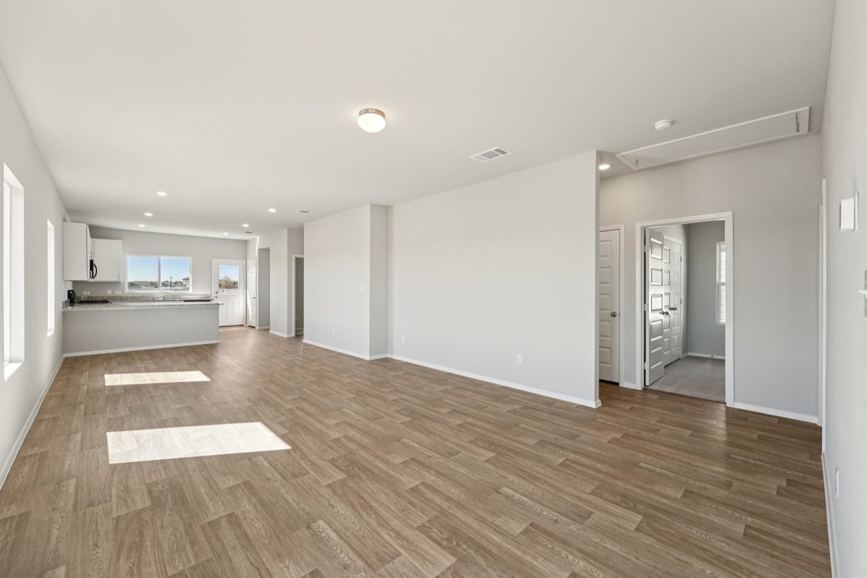 Image of a living area with light grey walls, vinyl flooring, white trim and a kitchen in the distance