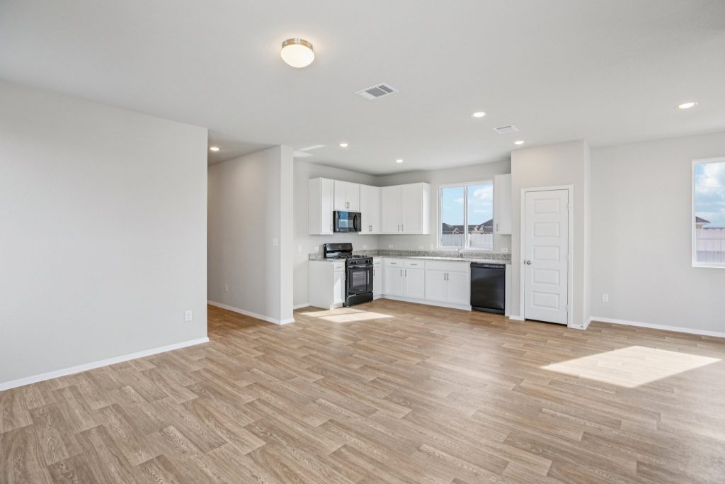 Image of a living room with an open kitchen with beige walls, light brown wood-look flooring and black appliances