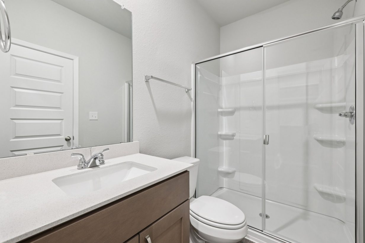 Image of a primary bathroom with brown cabinets, white vanity, a large mirror and a walk-in closet