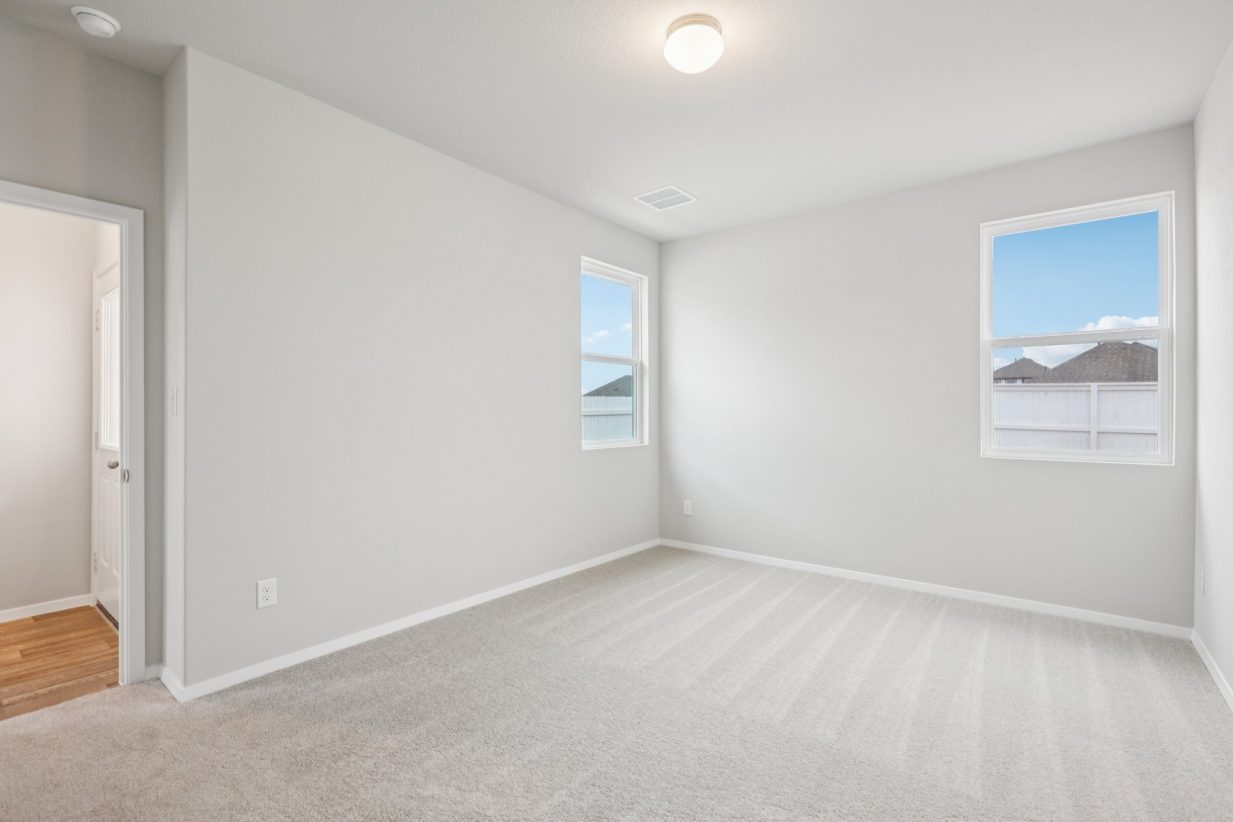 Image of a primary bedroom with light grey walls, tan carpeting, windows and white trim