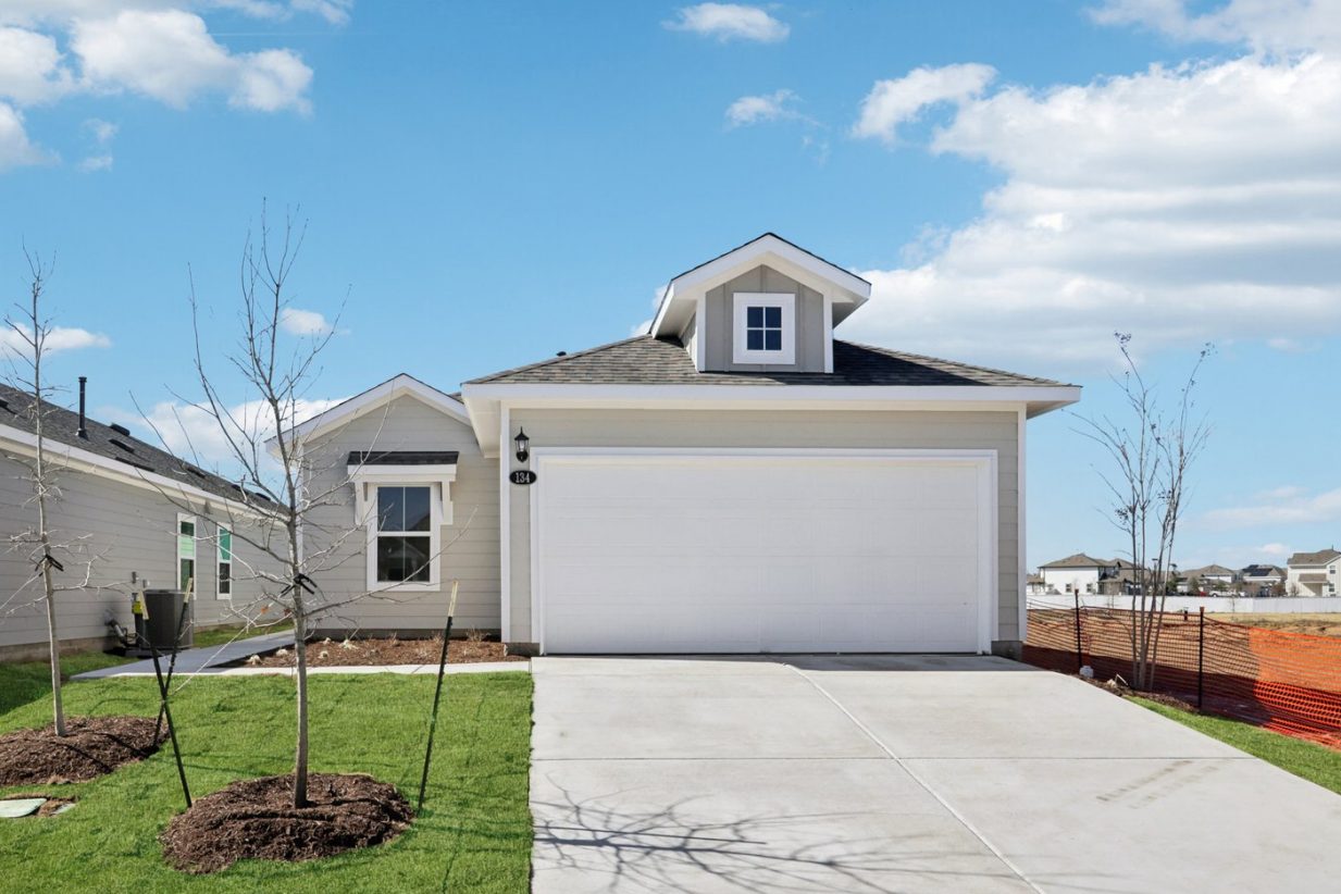 Image of the front exterior of a grey one story house with a white garage door, a cement driveway, a green front yard and a blue sky in the background