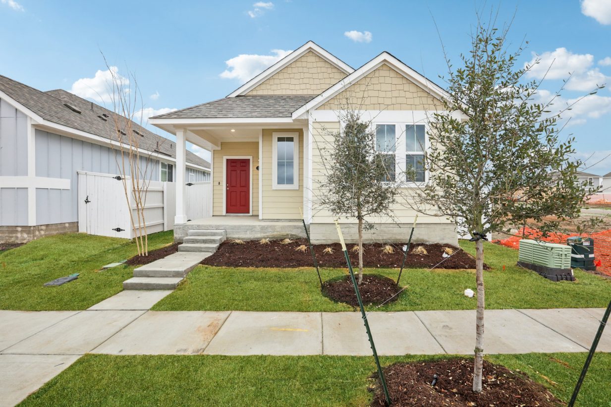 Image of a yellow one story house with a red front door, a green grass front yard, and a blue sky in the baclground