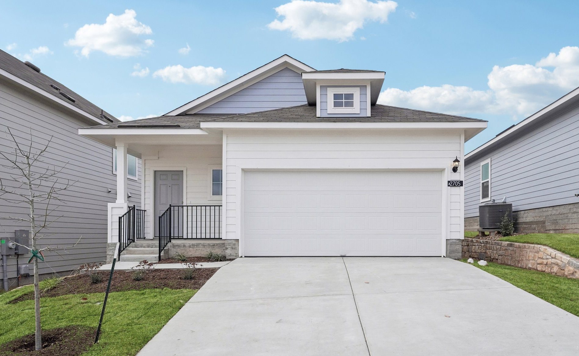 Image of a blue and white one story house with a white garage, a cement driveway, a green grass front yard and a blue sky in the background
