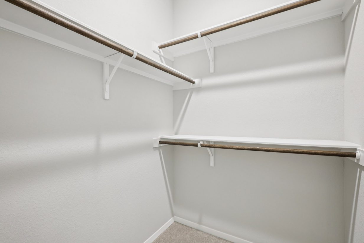 Image of a primary closet with light grey walls, tan carpeting and wooden rods