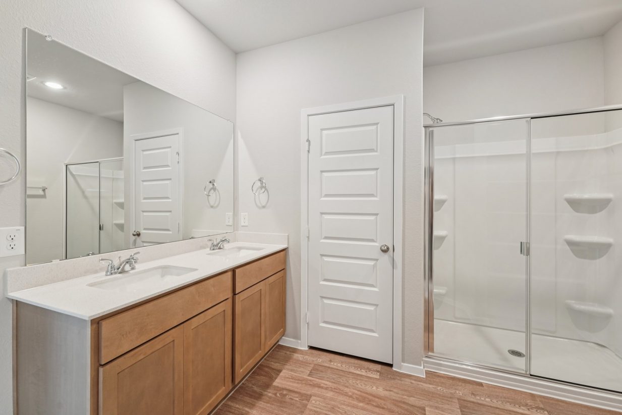 Image of a primary bathroom with light grey walls, a jack and jill white vanity, brown cabinets, a large mirror and a walk-in shower